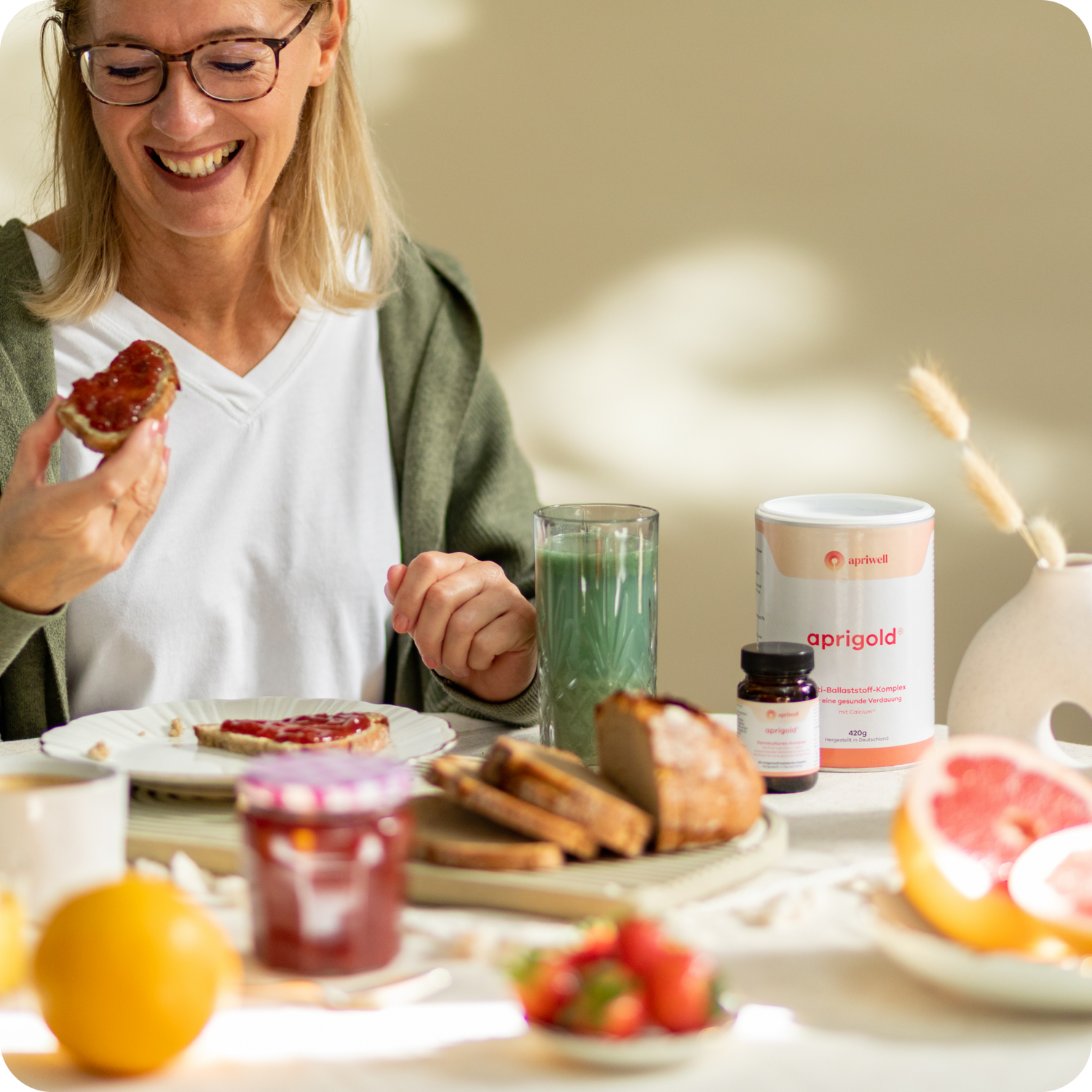 Eine Frau mit Brille lächelt und isst ein Stück Brot mit Marmelade. Auf dem Tisch stehen eine Dose aprigold Multi-Ballaststoff-Komplex, eine kleine Flasche Darmkulturen-Komplex, ein Glas mit einem grünen Getränk, Brot, Beeren, eine Orange und eine Grapefruit. Bildtyp: Produktbild