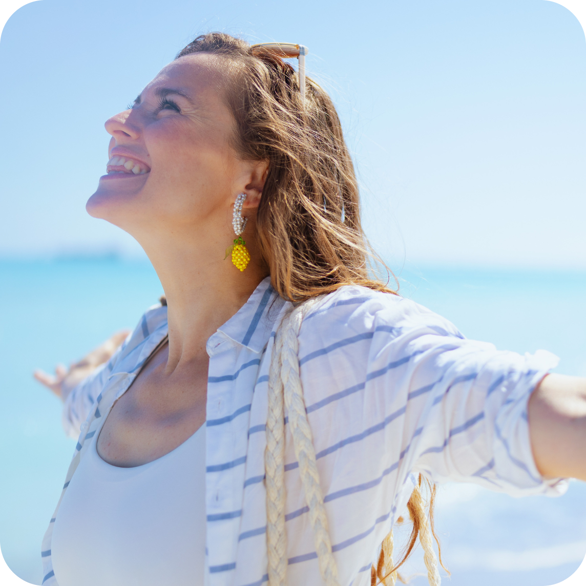 Eine Frau mit offenen Armen und lächelndem Gesicht blickt zum Himmel. Ihr Haar ist vom Wind zerzaust. Sie trägt eine weiße Bluse mit blauen Streifen und auffällige Ohrringe in Form von gelben Ananassen. Im Hintergrund ist das Meer und der Himmel zu sehen.