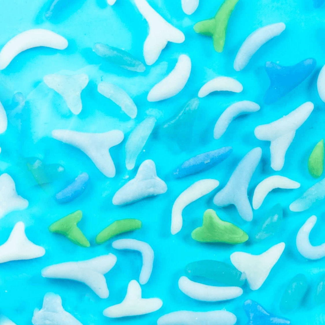 Produktbild – Nahaufnahme von bunten Zuckerstreuseln in verschiedenen Formen auf blauem Fondant, darunter mehrere in Form von Haifischflossen.