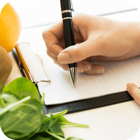 Beratung – zeigt eine Person, die etwas aufschreibt, umgeben von gesunden Lebensmitteln wie Spinat, Kiwi und Orange.
