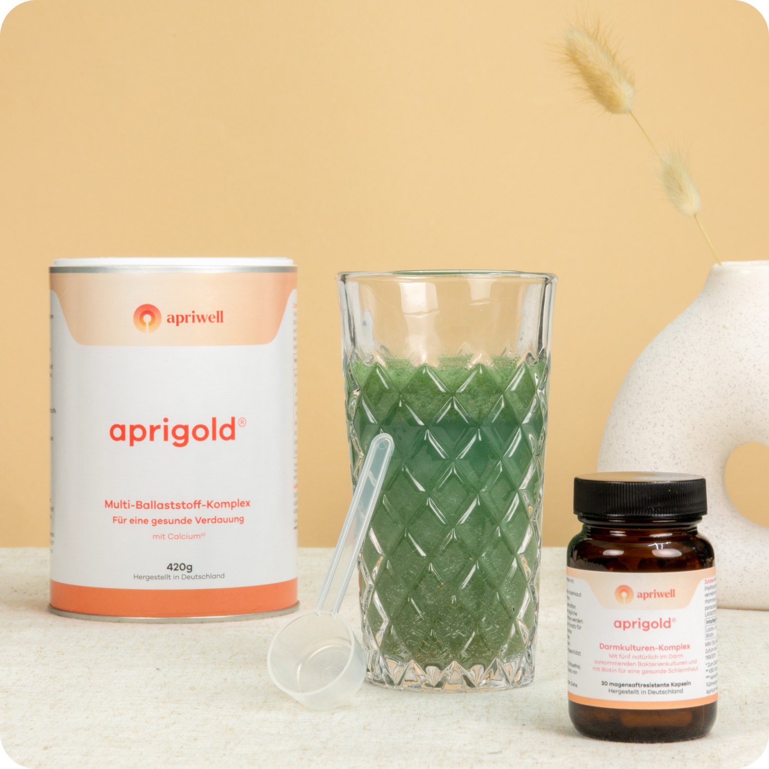 Produktbild: Eine Dose Aprigold Multi-Ballaststoff-Komplex mit Calcium, ein Glas mit grünem Getränk und ein Messlöffel sowie eine Braunglasflasche mit Aprigold Darmkulturen-Komplex.