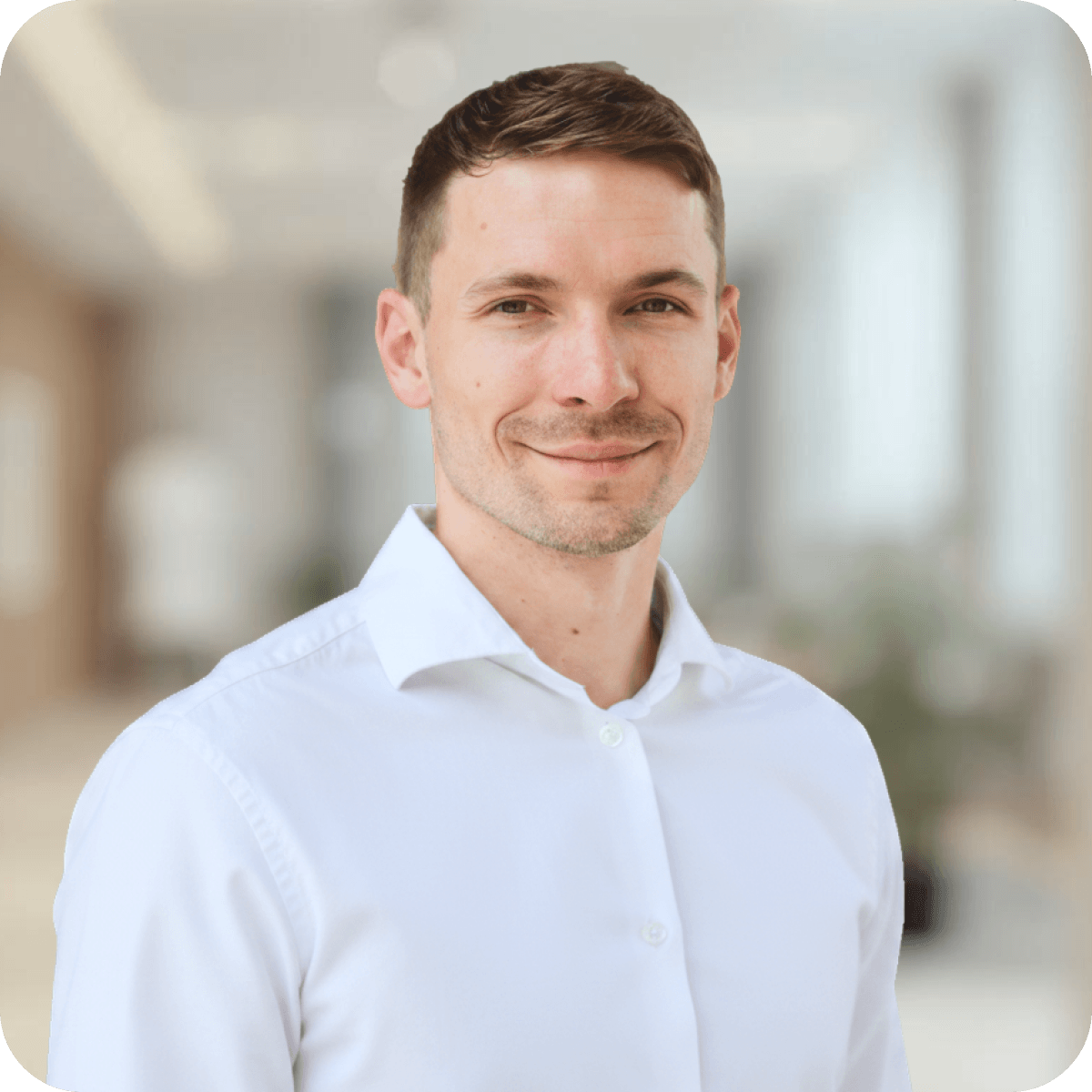 Beratung: Erik, Ansprechpartner, blickt freundlich lächelnd in die Kamera. Er trägt ein weißes Hemd vor einem hellen, unscharf gestalteten Hintergrund in einer Büroumgebung.