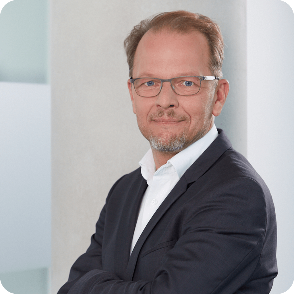 Ein lächelnder Mann mit Brille, Bart und dunkelblondem Haar, der einen dunklen Anzug und ein weißes Hemd trägt und die Arme verschränkt hat.